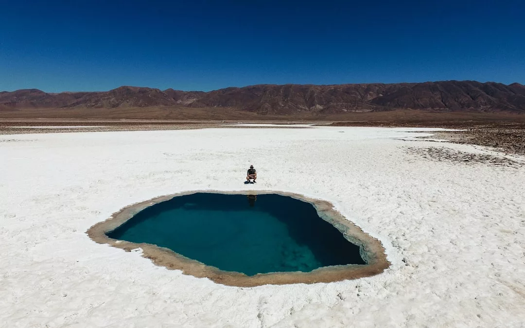 A person sits at the edge of a deep blue pool surrounded by white salt flats, with mountains and a clear blue sky in the background—offering an idyllic scene perfect for unique real estate opportunities.
