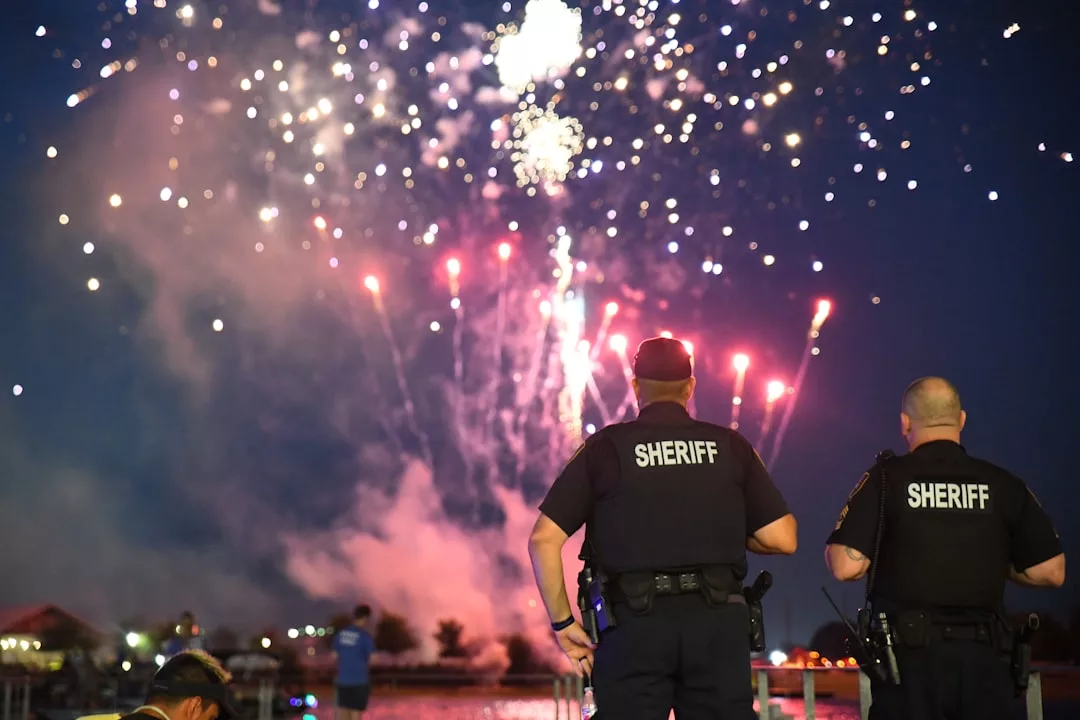 Two sheriff officers stand facing a night sky filled with fireworks, observing the event as people near a body of water enjoy the celebration—an inviting scene that highlights community spirit and local real estate charm.