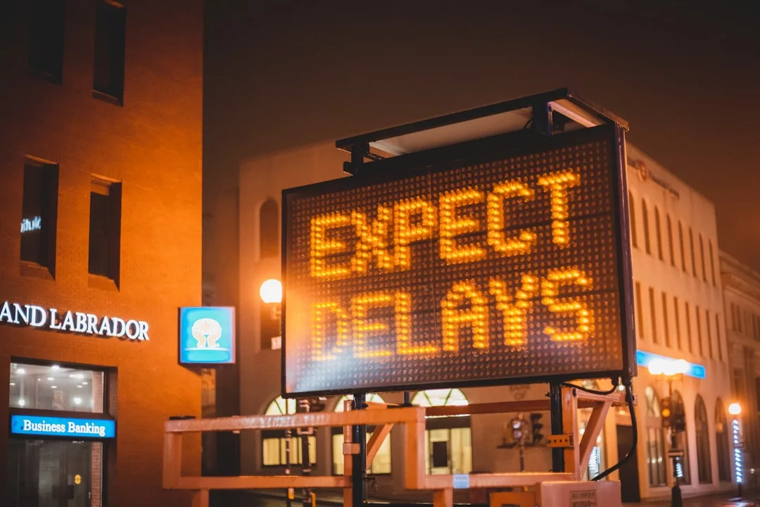 Electronic road sign displays "EXPECT DELAYS" in bright lights on a city street at night, with buildings and real estate properties visible in the background.