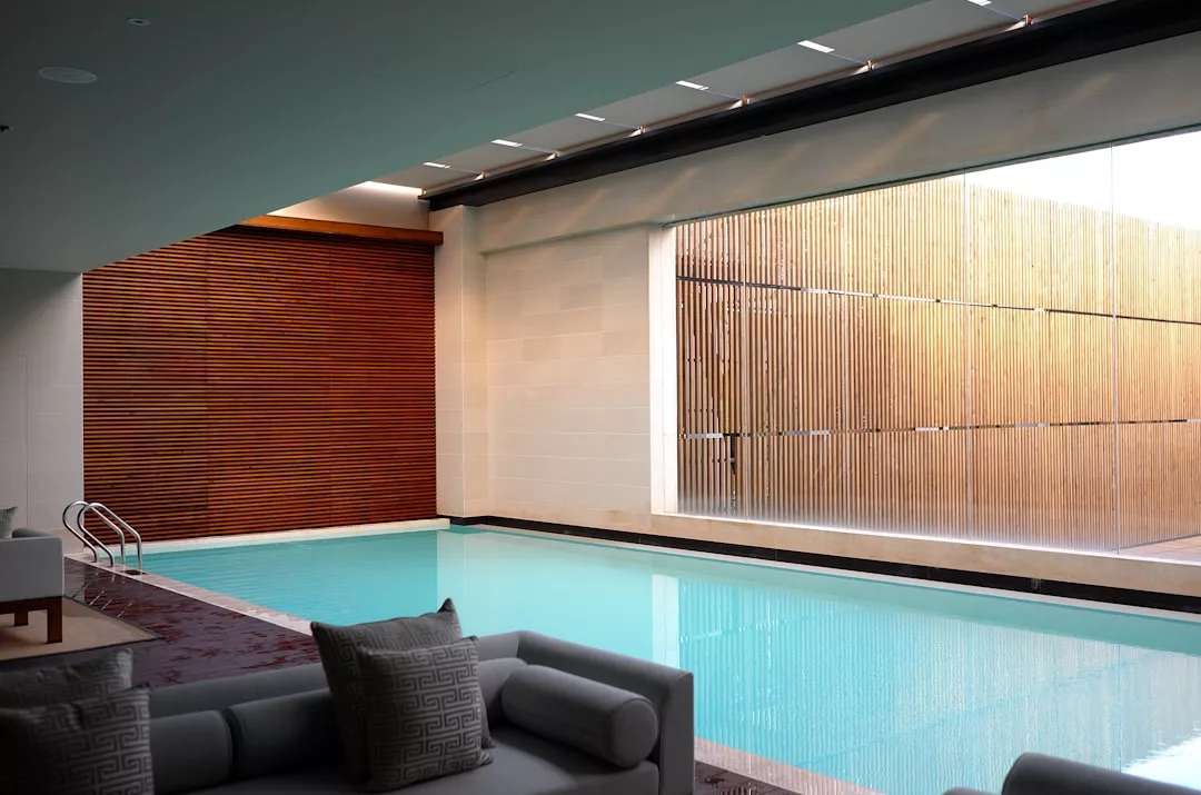 Indoor swimming pool with clear blue water, surrounded by modern seating—perfect for real estate buyers. Large windows with wooden slat coverings allow natural light to fill this inviting space.
