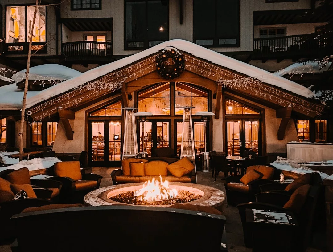 Cozy outdoor seating with cushioned chairs surrounds a fire pit in front of a lodge-style building, beautifully lit and covered in snow at night—a dream Real Estate retreat for winter evenings.