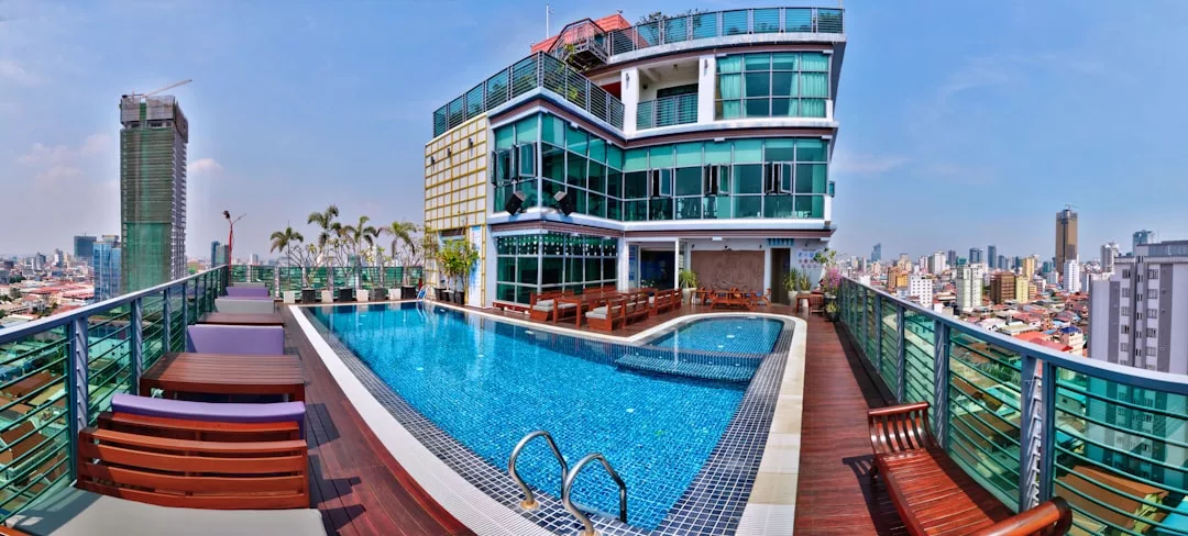 Rooftop swimming pool surrounded by lounge chairs, with a modern glass building and city skyline in the background under a clear sky—an exceptional real estate offering for urban luxury living.