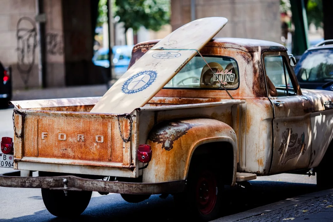 A rusty vintage Ford pickup truck with a surfboard sticking out of its bed is parked on a city street, capturing the laid-back vibe that makes this area’s real estate so unique and appealing.