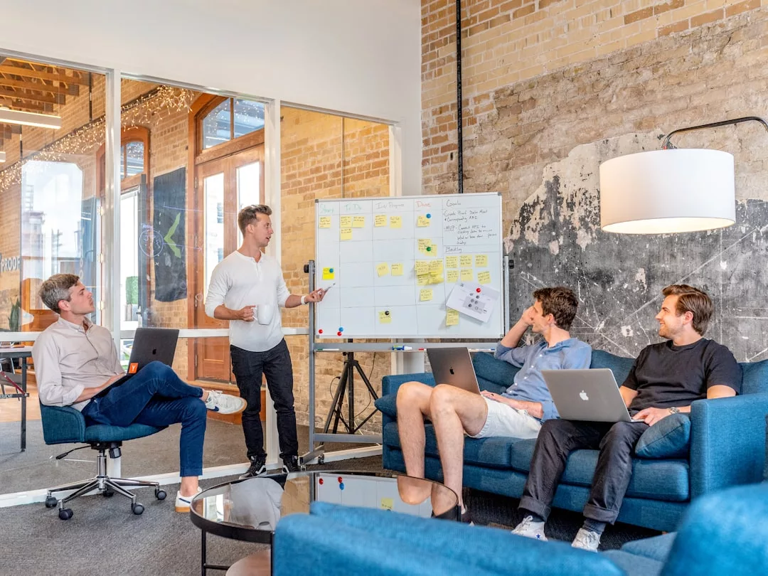 Four men in a modern office sit and listen as one stands, presenting real estate strategies on a whiteboard filled with sticky notes and diagrams.