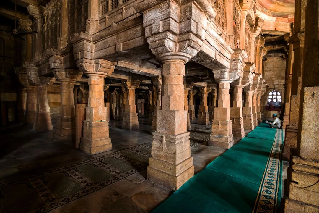 Interior view of a historic stone mosque, ideal for unique real estate opportunities, featuring multiple columns, a patterned floor, green prayer rugs, and a person sitting near the window.