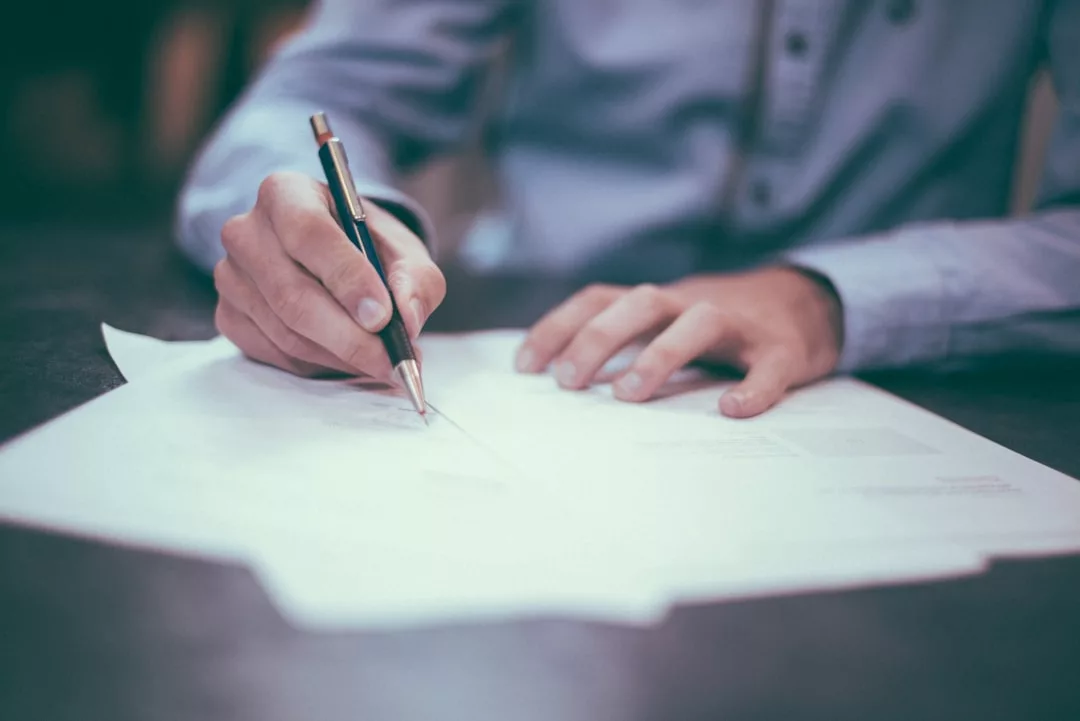 A person in a blue shirt writes on a stack of real estate papers with a pen at a desk.