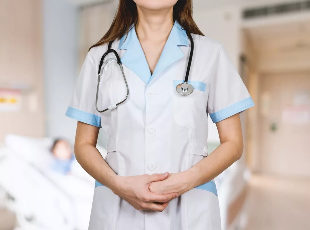 A medical professional in a white coat with a stethoscope stands with hands clasped in a hospital room, reminiscent of the precision and care seen in real estate—where every detail matters; a patient bed is visible in the background.