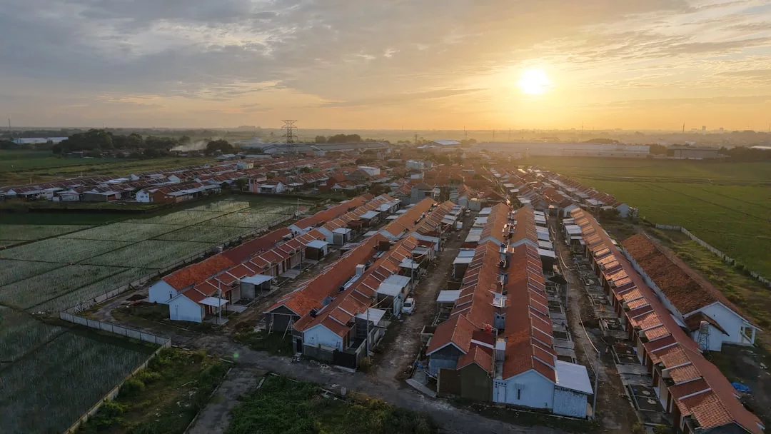 Aerial view of a residential neighborhood with rows of red-roofed houses, surrounded by green fields under a sunrise sky—a picturesque real estate landscape.