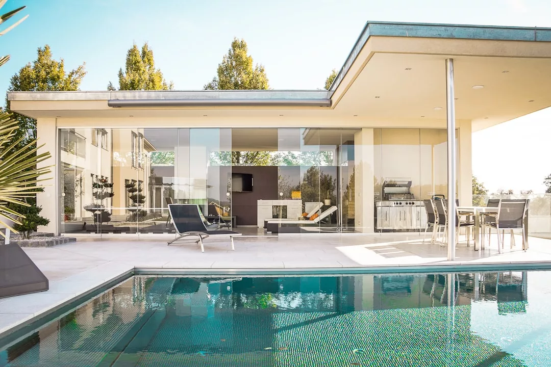 Modern real estate features large glass windows, outdoor lounge chairs, a dining area, and a swimming pool in the foreground under a clear sky.