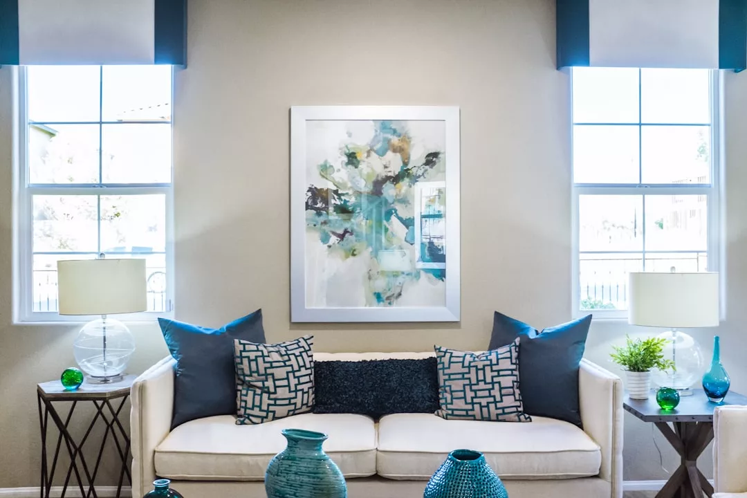 A modern living room perfect for real estate showcase, featuring a white sofa, blue and patterned pillows, abstract wall art, side tables with lamps, decorative blue and green vases, and large windows that fill the space with natural light.