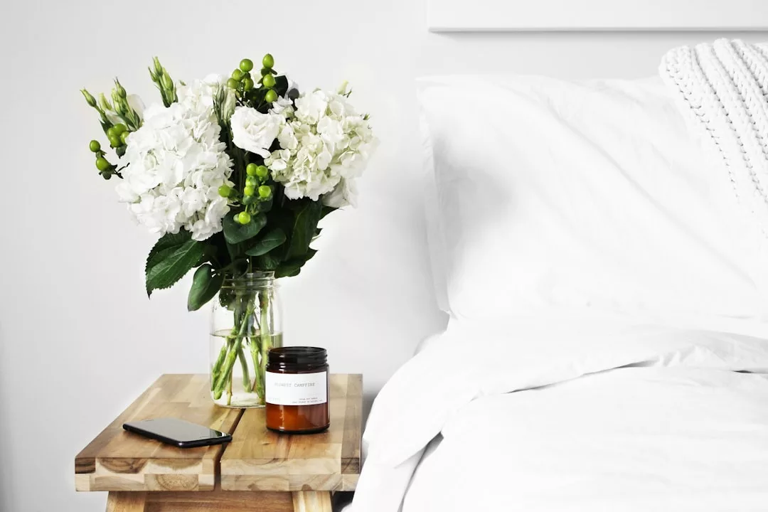 A wooden bedside table holds a jar candle and a smartphone—an inviting feature often highlighted in real estate listings—while a glass vase of white flowers sits next to a neatly made white bed.