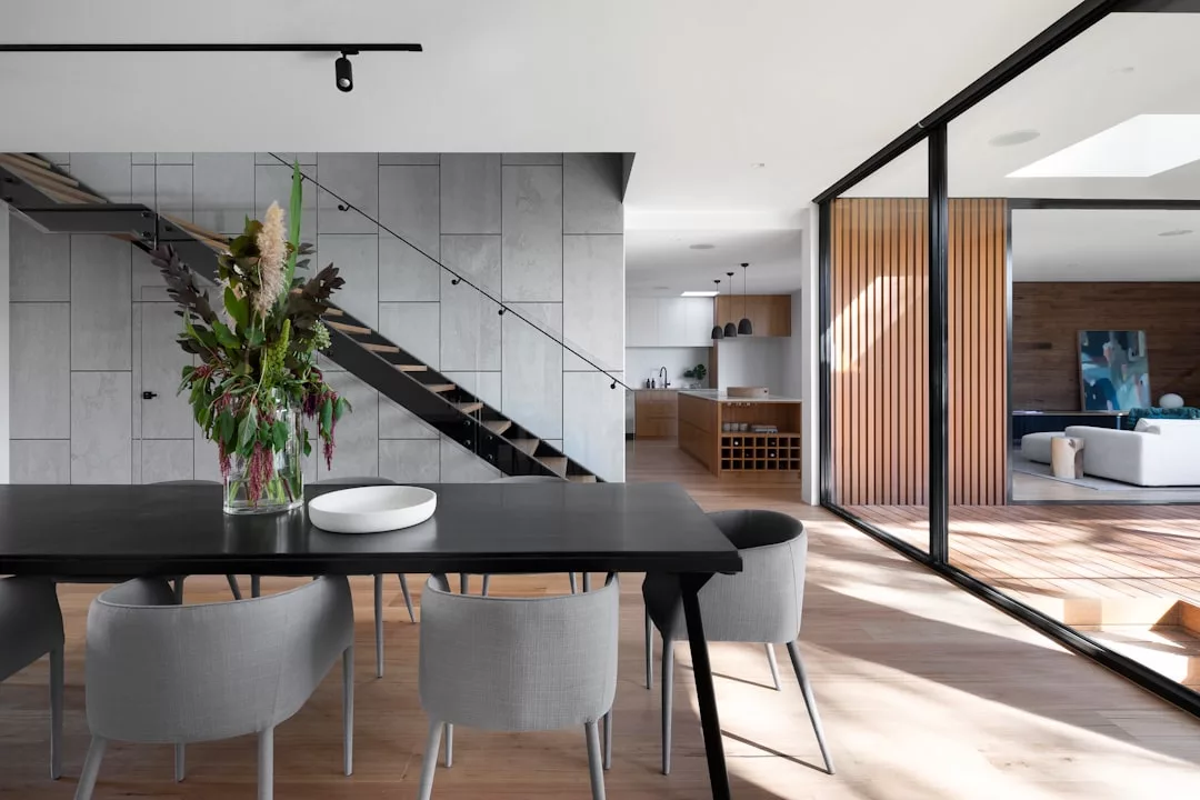 Modern dining area with a black table, gray chairs, and a large floral arrangement, adjacent to an open living space with wood accents and large sliding glass doors—perfect for stylish real estate showings.