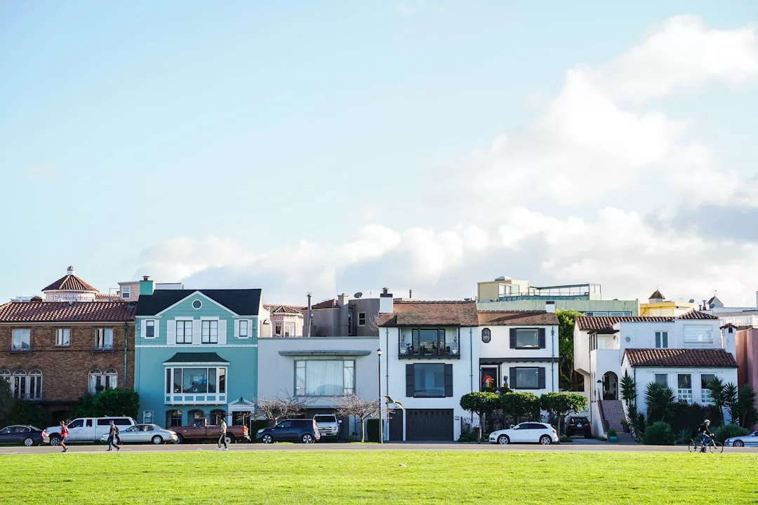 Row of colorful houses with cars parked on the street and people walking or cycling in front reflects suburban living near Stockholm outskirts, with a large grassy area and blue sky in the background.