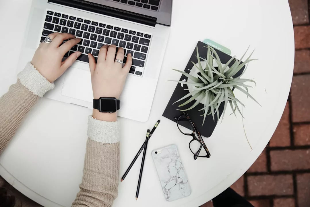 Person typing on a laptop at a round white table, working on real estate notes with a notebook, plant, smartphone, eyeglasses, and two pencils.