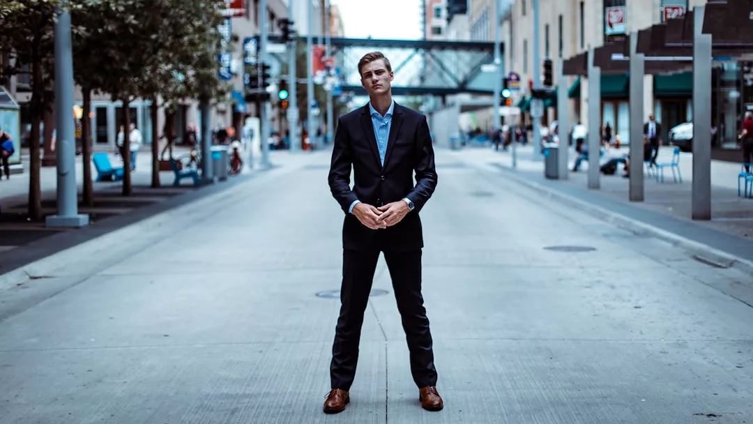 A man in a dark suit stands in the middle of an empty city street, facing the camera, with buildings and a skywalk in the background—capturing the essence of urban real estate.