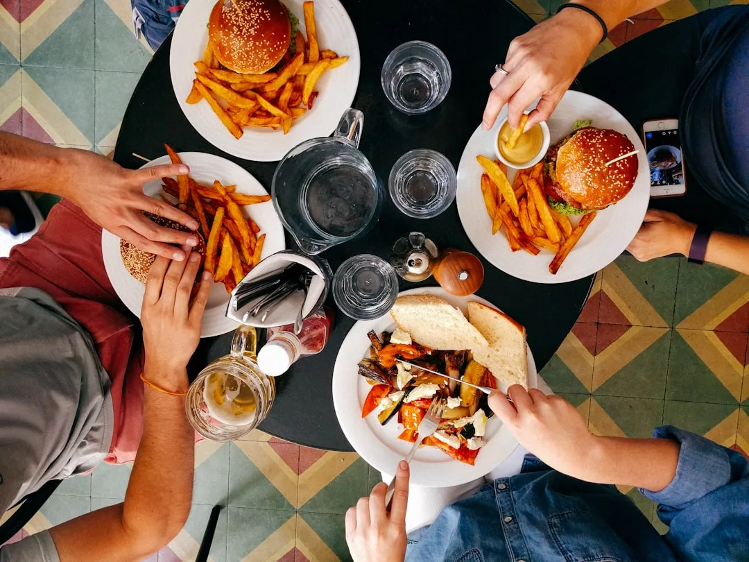Three people sit at a round table in a modern real estate setting, eating burgers and fries with drinks and utensils laid out, photographed from above.
