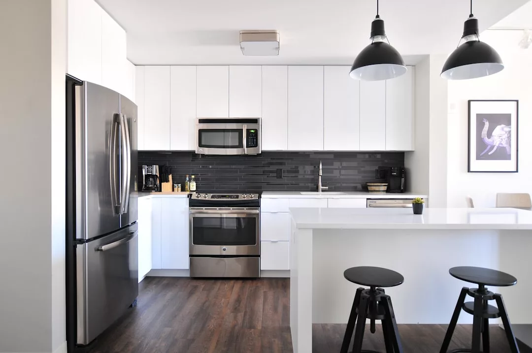 Modern kitchen with white cabinets, stainless steel appliances, black backsplash, and island with two black stools—ideal for those considering when to sell property in Copenhagen. Pendant lights and framed artwork add stylish touches.