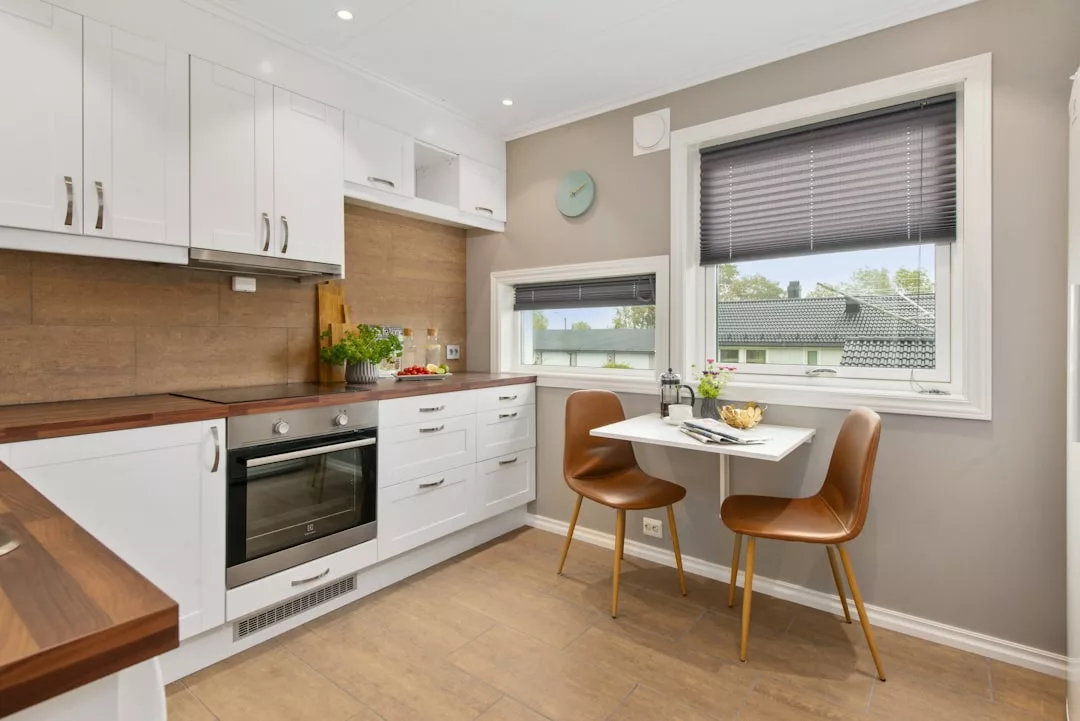 Modern kitchen with white cabinets, wooden countertops, built-in oven, and a small table set for two by a window with brown blinds—perfect for real estate buyers seeking style and comfort.