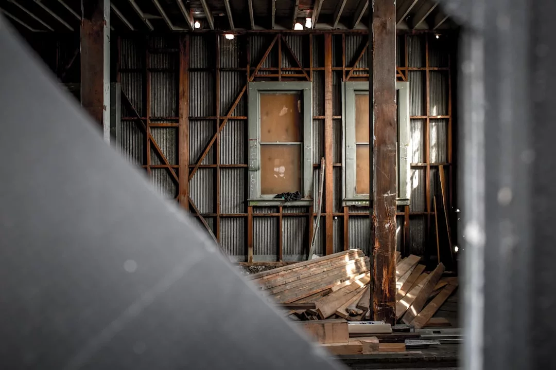 Exposed wooden beams and unfinished walls with two boarded windows and scattered lumber create a raw atmosphere in this partially constructed or abandoned real estate property.