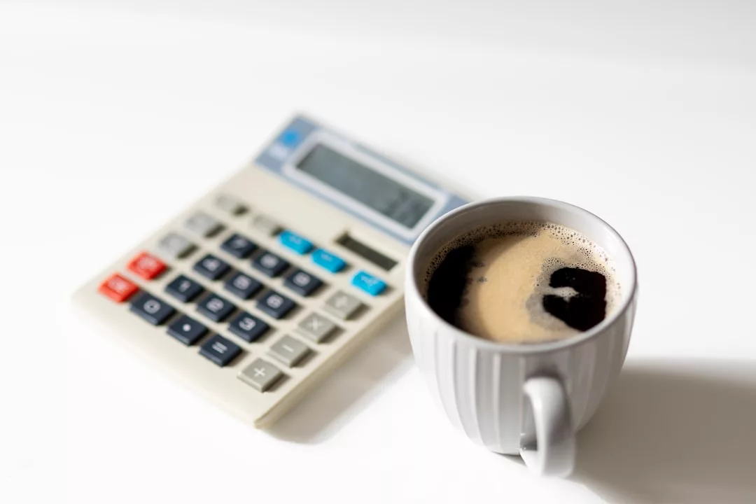 A white coffee cup filled with black coffee sits in front of a calculator on a white surface, creating the perfect setting for tackling early morning real estate calculations.
