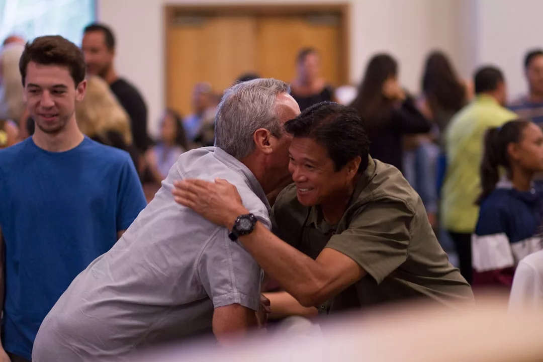 Two men greet each other with a hug and a handshake in a crowded real estate event, while others stand and talk in the background.