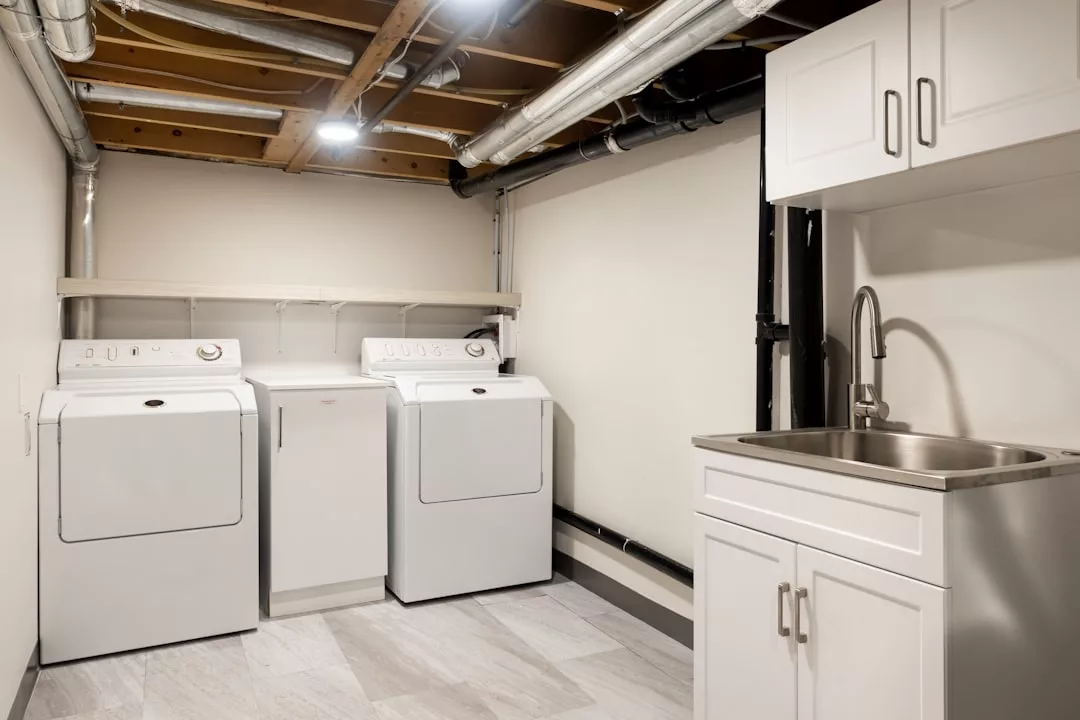 A laundry room perfect for real estate listings, featuring a washing machine, dryer, white cabinetry, stainless steel sink, exposed ceiling pipes, and a tiled floor.