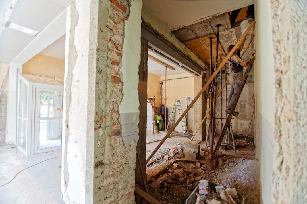 A construction worker stands on scaffolding inside a real estate property under renovation, with exposed brick walls, debris, and construction tools visible.