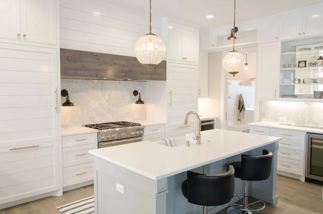Modern kitchen with white cabinets, a marble backsplash, stainless steel appliances, an island with a sink, two black chairs, and two pendant lights—perfect for real estate buyers seeking both style and functionality.