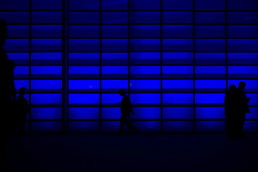 A person walks in front of a large blue-lit real estate wall, while two other silhouetted figures stand and look at their phones on the right side.