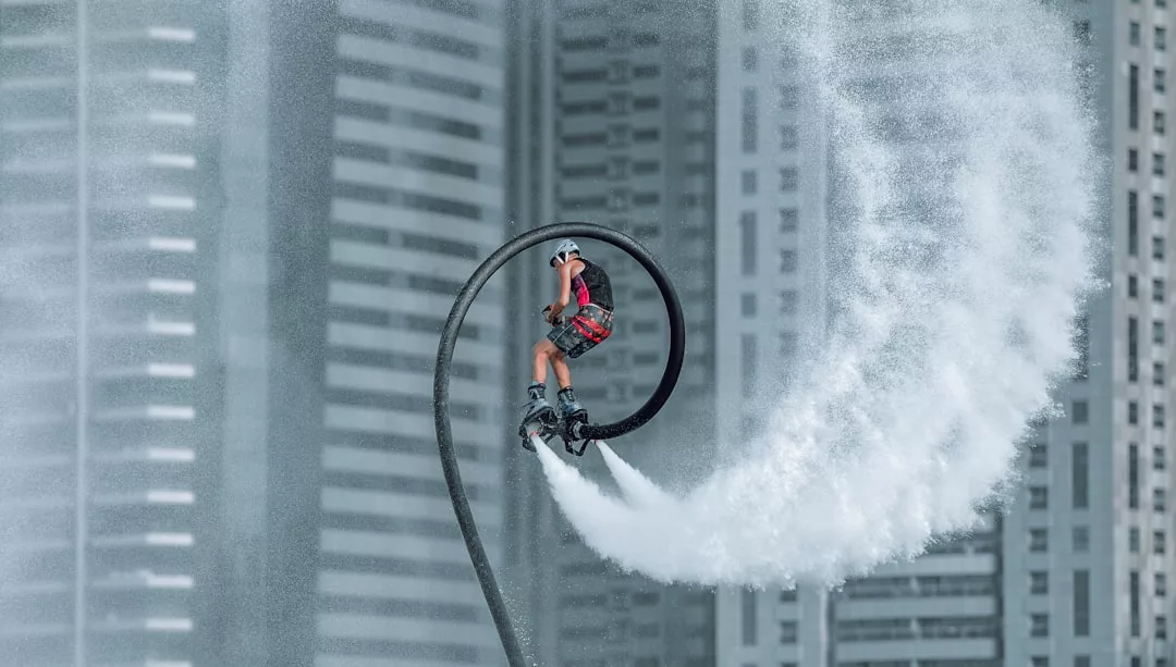 A person rides a water-powered flyboard high above the water, creating a spiral arc of spray, with impressive real estate and tall buildings in the background.