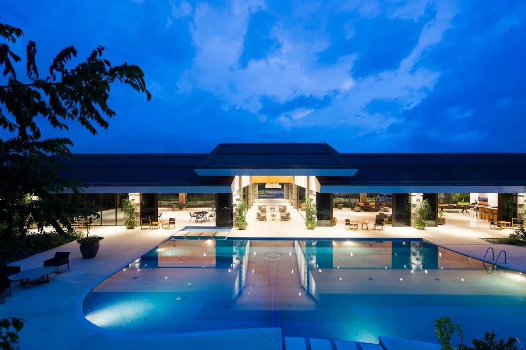 Modern Real Estate building with a large outdoor swimming pool, illuminated at dusk against a backdrop of blue sky and clouds.