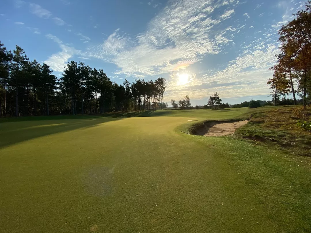 A golf course green with a sand bunker on the right, surrounded by trees—an ideal real estate opportunity—under a partly cloudy sky with the sun low on the horizon.
