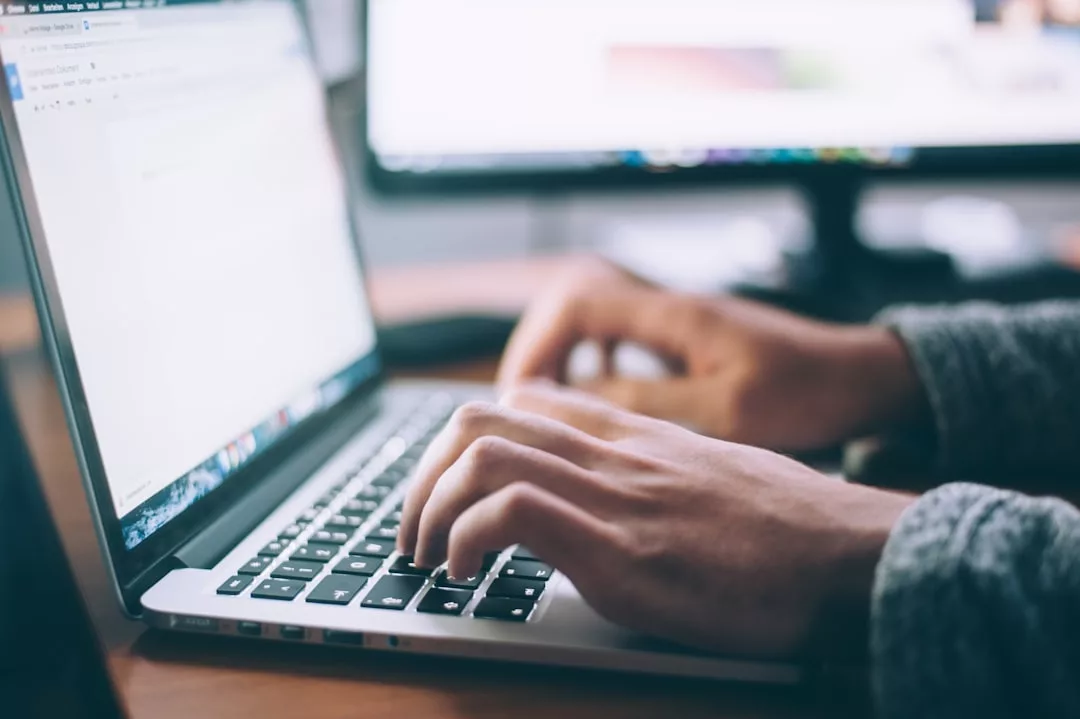 A person types on a laptop keyboard at a desk with a computer monitor in the background, researching real estate opportunities.
