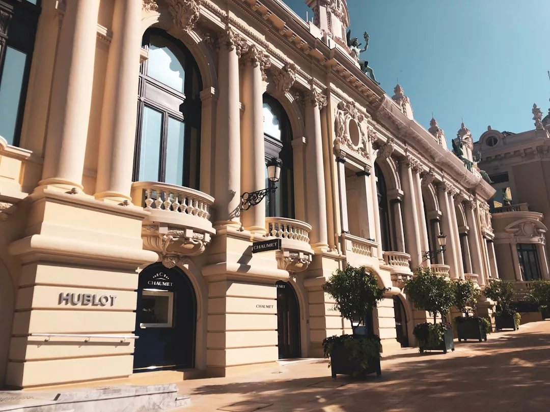 A grand beige stone building with ornate columns, arched windows, and small trees in planters lines the entrance under a clear blue sky—a stunning piece of real estate.