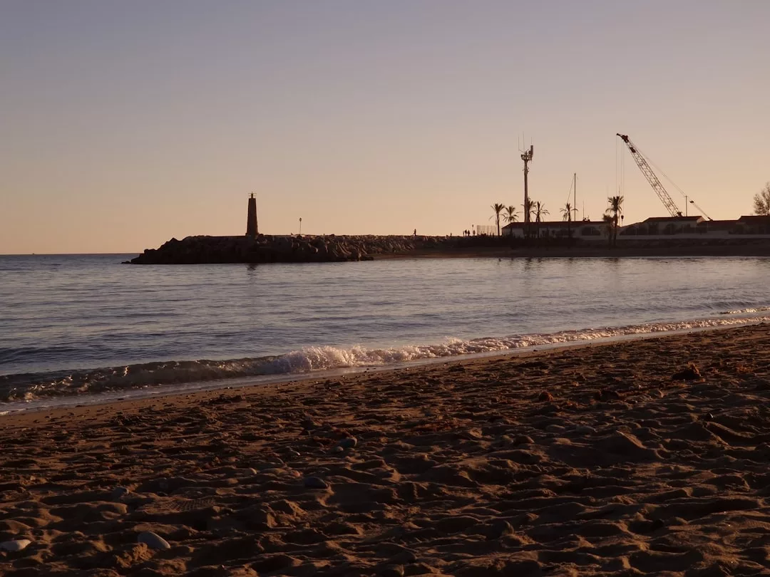 A sandy beach at sunset with calm waves, a rocky jetty, lighthouse, and construction crane hint at nearby real estate development, while palm trees sway gently in the background.