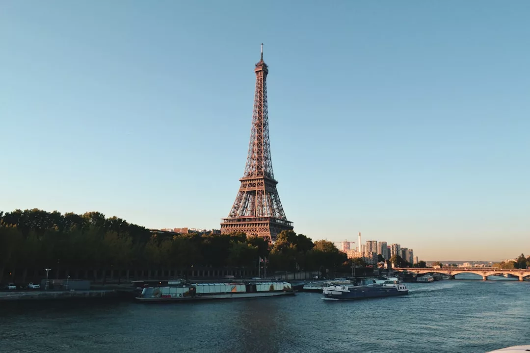 The Eiffel Tower is seen from across the Seine River, with boats docked along the riverbank and city buildings—including prime real estate—rising in the background under a clear sky.
