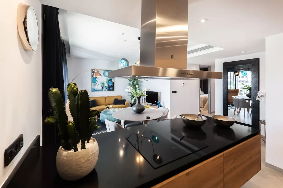 Modern open-plan kitchen with a black countertop, potted cactus, stovetop, and range hood, overlooking a living area with a yellow sofa and dining table—ideal for stylish real estate living.