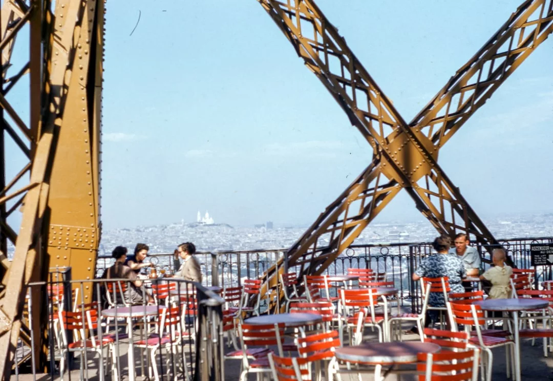 People sit at red and white tables on an outdoor terrace framed by the Eiffel Tower’s metal beams, enjoying a view of Paris real estate and the city skyline in the background.