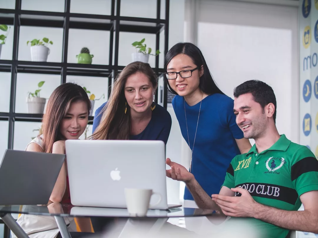 Four people are gathered around a table looking at a laptop screen, engaged in a real estate discussion in a modern office setting with plants on shelves in the background.