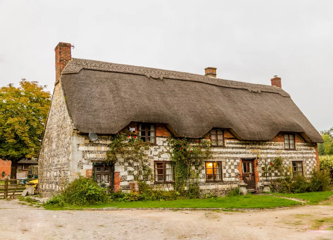 A charming stone cottage with a thatched roof, ivy-clad walls, and chimneys at each end sits in a grassy yard under cloudy skies—a picturesque real estate gem.