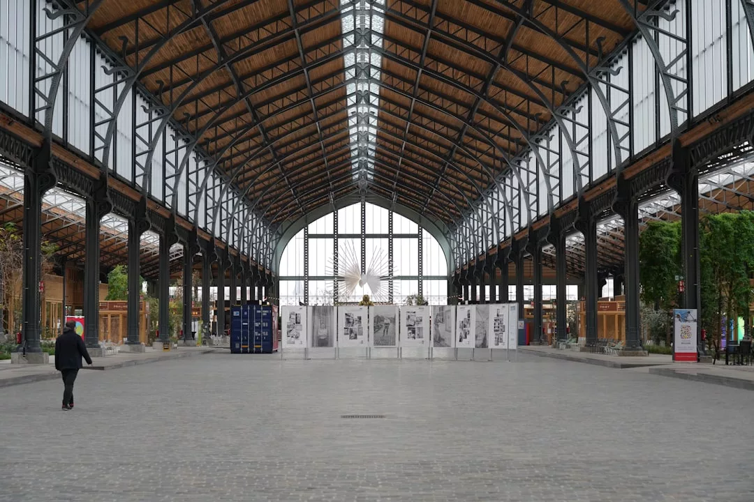 Spacious interior of a large, open hall with a high wooden roof, glass walls, and metal columns; ideal for Real Estate showcases. A person walks on the left, and temporary partitions stand at the far end.