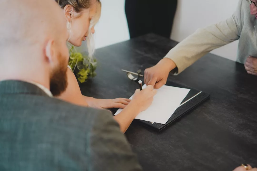 Three people sitting at a table, with one person signing a real estate document while another points at the paper.
