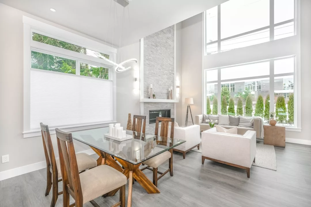 Modern open-plan living and dining area perfect for real estate showcases, featuring a glass table, wooden chairs, white armchairs, fireplace, large windows, and abundant natural light.