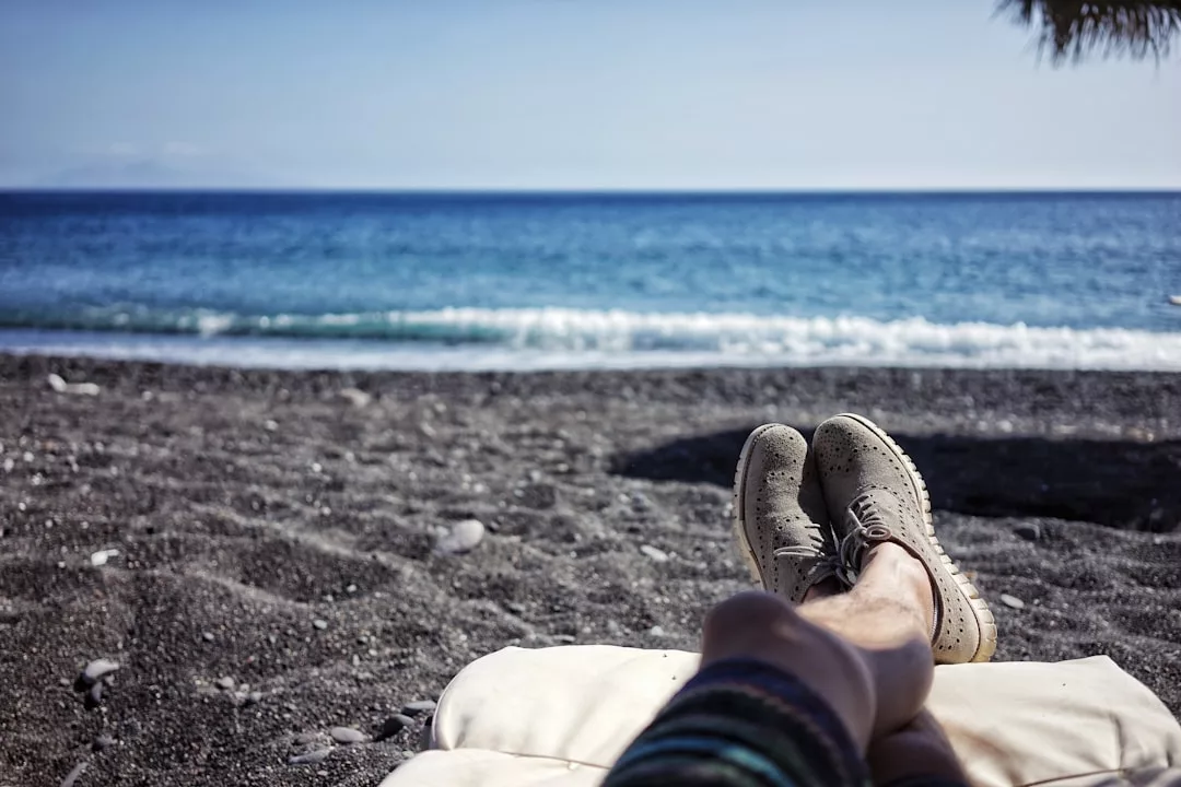 Person lying on a lounge chair at a black sand beach with legs extended, facing the ocean under a clear blue sky—an idyllic scene highlighting the serenity offered by coastal real estate.