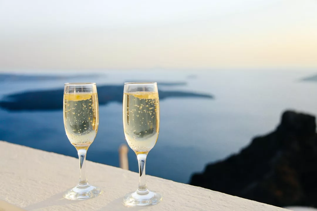 Two glasses of sparkling wine on a white ledge overlook a calm sea and distant islands at sunset, capturing the tranquil allure of coastal real estate.