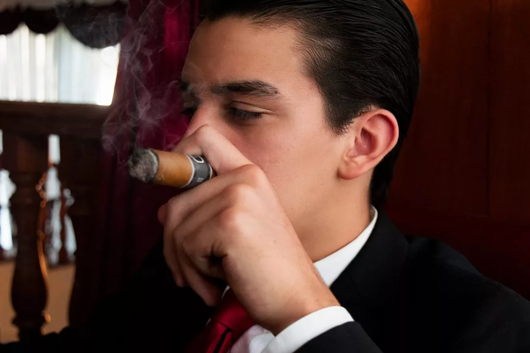 A man in a suit and red tie, exuding real estate power, smokes a cigar indoors as smoke curls near his face.