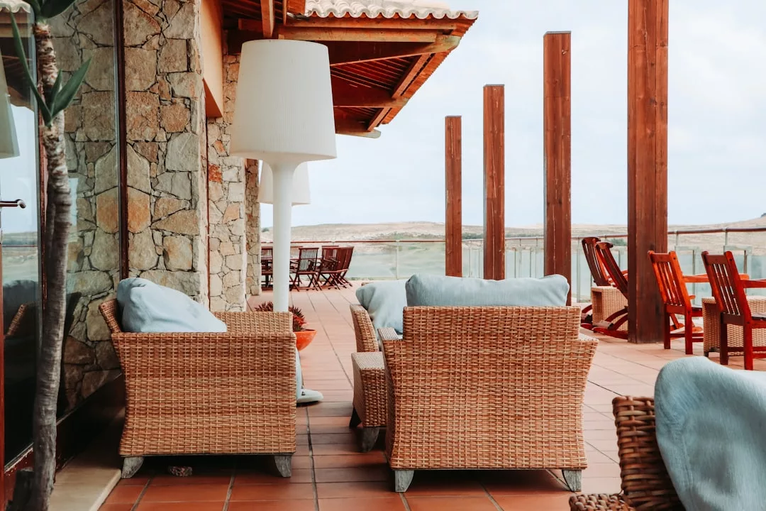 Wicker chairs with cushions and a floor lamp on a tiled patio, featuring wooden beams and outdoor dining furniture in the background—perfect for enjoying your Real Estate’s inviting outdoor space.