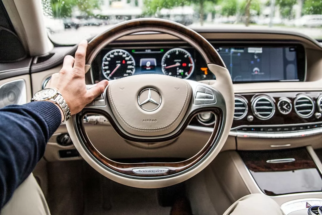 A person’s left hand is on the steering wheel of a Mercedes-Benz car, with the dashboard, gauges, and infotainment screens visible—capturing a sleek ride to a real estate showing.