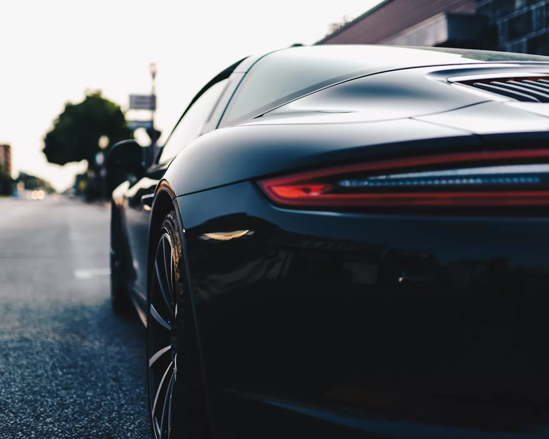 Close-up rear view of a sleek black sports car parked on a city street near modern real estate, showcasing the taillight, curved body, and rear wheel in focus.