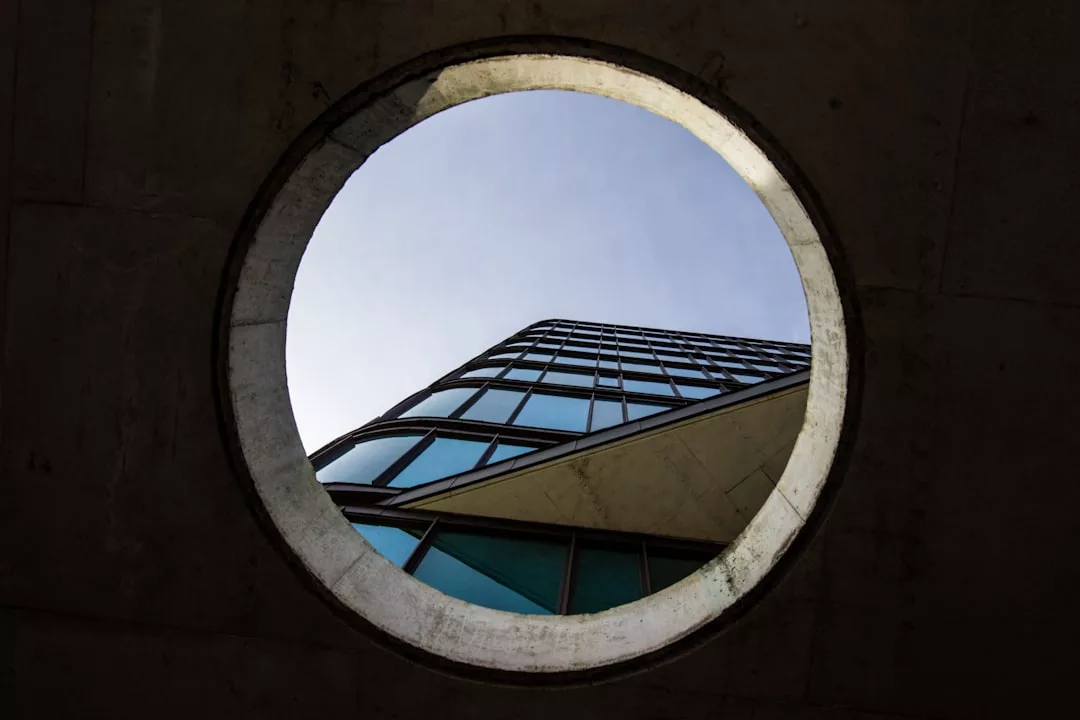 View of a modern glass building seen through a large circular opening in a concrete structure, highlighting the allure of Luxury Properties Copenhagen against the sky—a testament to stable investment in contemporary architecture.